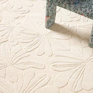 Puede incluir: Alfombra color crema con un gran estampado floral. El dise&ntilde;o de la alfombra presenta formas de flores estilizadas en relieve. Una silla con un estampado floral en el asiento y las patas es parcialmente visible, a&ntilde;adiendo contraste.