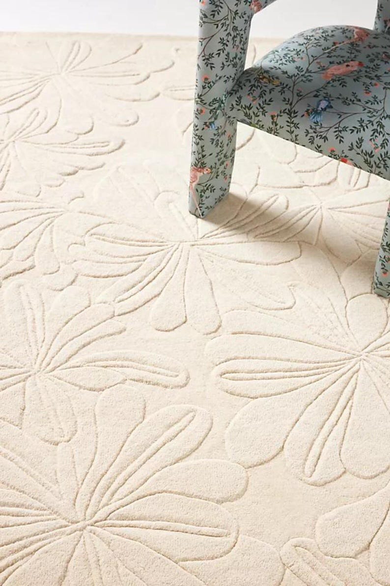 Puede incluir: Alfombra color crema con un gran estampado floral. El dise&ntilde;o de la alfombra presenta formas de flores estilizadas en relieve. Una silla con un estampado floral en el asiento y las patas es parcialmente visible, a&ntilde;adiendo contraste.