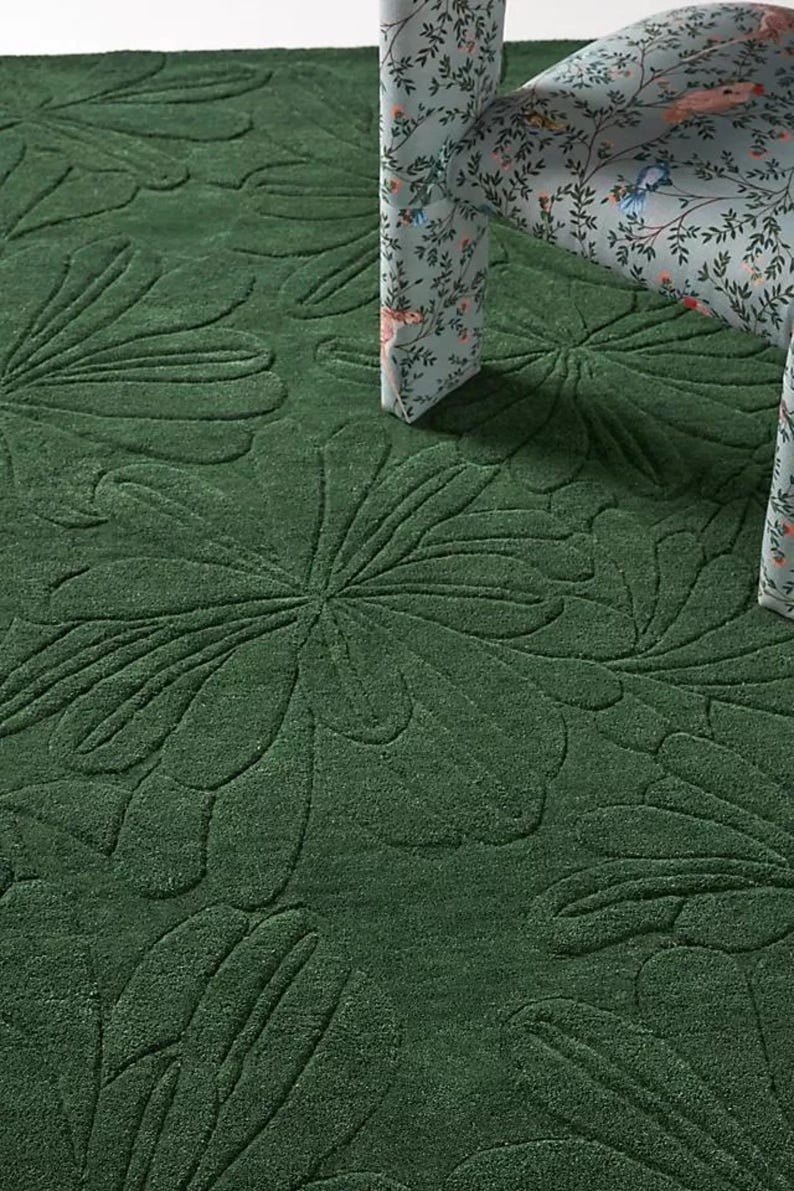 May include: A dark green rug with a raised floral pattern. The rug's texture is visible, and a chair with a floral pattern is partially visible in the background.