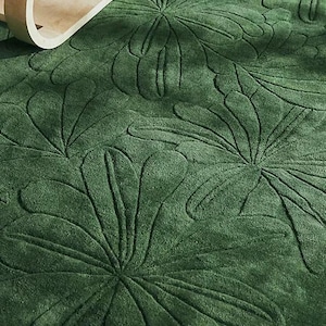 May include: A close-up of a green rug with a floral pattern. The rug has a textured surface with raised floral designs. A wooden side table with books and a bowl is in the background.