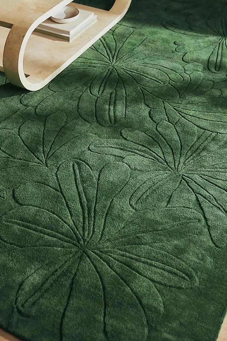 May include: A close-up of a green rug with a floral pattern. The rug has a textured surface with raised floral designs. A wooden side table with books and a bowl is in the background.