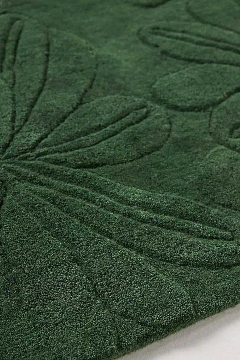 May include: Close-up of a dark green rug with a floral leaf pattern. The design is embossed, creating a textured surface. The rug's color is a deep, rich shade of green, suitable for home decor.