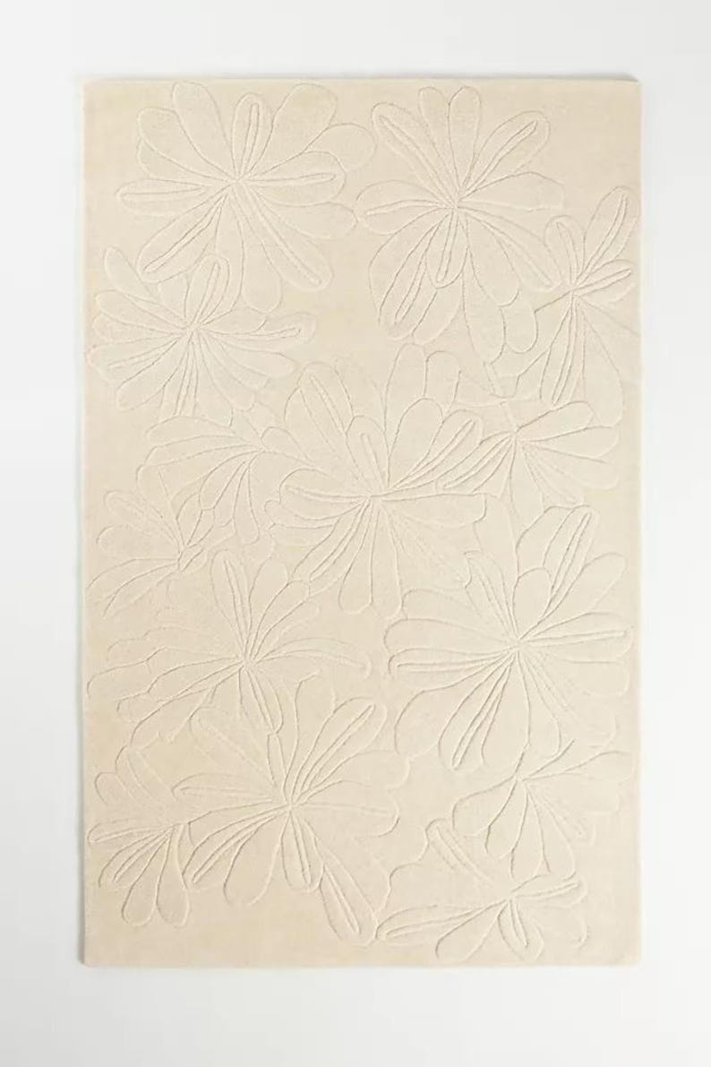 Puede incluir: Una alfombra rectangular de color crema con un patr&oacute;n floral repetitivo. Las flores est&aacute;n estilizadas con m&uacute;ltiples p&eacute;talos y est&aacute;n en relieve, creando un efecto texturizado. La alfombra est&aacute; sobre un fondo blanco.