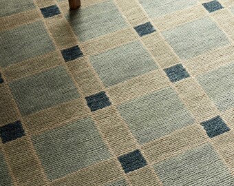 May include: A close-up of a patterned rug with a grid design. The rug features squares of light blue and beige, with dark blue accent squares. The texture of the rug is visible, and a wooden chair leg is in the upper left corner.