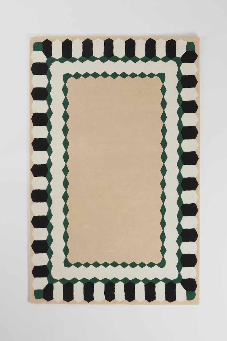 May include: Rectangular rug with a beige center and a patterned border. The border features alternating black and white geometric shapes, with a green diamond pattern in between. The rug is designed for home decor.