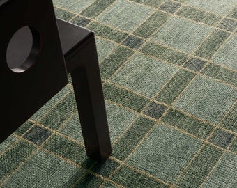 May include: A close-up of a green and beige plaid rug with a black chair. The rug has a textured appearance with a grid-like pattern. The chair is black and has a circular cutout in the backrest. The rug's design is a classic plaid.