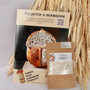 Op de afbeelding: Een bruine papieren zak met een wit label waarop "See Fania Sourdough Starter" staat en wit poeder erin. De zak ligt naast een kookboek met een brood op de omslag. Het kookboek heeft de titel "Recepten met zuurdesem" en bevat Oekraïense tekst.
