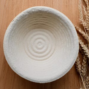 Op de afbeelding: Een ronde, gebroken witte rijsmand met concentrische cirkelpatronen. De mand staat op een houten oppervlak, met een bosje tarwearen rechts. Geschikt voor ambachtelijk brood bakken.