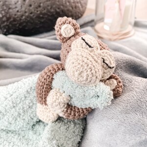 Op de afbeelding: Een gehaakt aapje met bruin en beige bont, dat een lichtblauw dekentje vasthoudt. De aap ligt op een grijs dekentje.