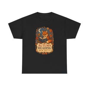 Könnte beinhalten: Ein schwarzes T-Shirt mit einer zentralen Grafik eines Fuchses, der ein Buch liest, umgeben von Herbstblättern, einer Mondsichel und Sternen. Unter dem Fuchs befindet sich ein dekorativer Rahmen mit dem Text "AUTUMN READING SOCIETY".