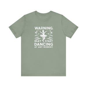 Puede incluir: Camiseta verde salvia con un gráfico blanco de una bailarina y el texto "ADVERTENCIA: PUEDE EMPEZAR A BAILAR EN CUALQUIER MOMENTO."