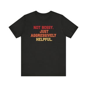May include: Black t-shirt with the text "NOT BOSSY. JUST AGGRESSIVELY HELPFUL." in red, orange, and yellow lettering. The text has a distressed, vintage look. The shirt is made of a soft material.