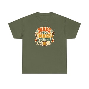 Könnte beinhalten: Ein olivgrünes T-Shirt mit einem zentralen Aufdruck, der den Text "MADE TO MAKE THINGS" in Orange und Petrol zeigt. Das Design enthält Illustrationen von einer Schere, einem Pinsel und einer Garnrolle, akzentuiert durch kleine orangefarbene Blumen und Sterne.