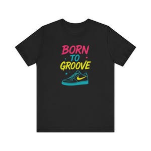 Puede incluir: Camiseta negra con el texto "BORN TO GROOVE" en rosa y amarillo, y un gráfico de una zapatilla deportiva con un swoosh amarillo. La zapatilla está delineada en azul.