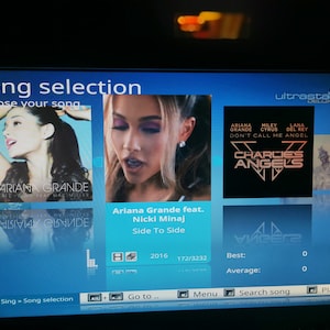 May include: A screen displaying a song selection menu with a blue background. The song "Side To Side" by Ariana Grande featuring Nicki Minaj is highlighted. The song is from 2016 and is 172 seconds long out of a total of 3232 seconds. The screen also shows a playlist option and a search song option.