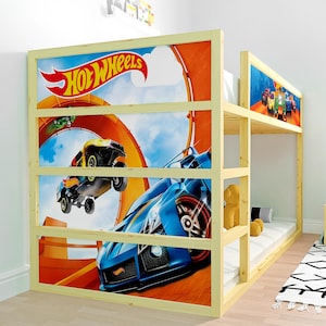 May include: A wooden bunk bed with a Hot Wheels theme. The headboard and side panel feature a vibrant graphic of race cars on a track. The bed frame is made of light-colored wood, and the design includes a lower and upper bunk.