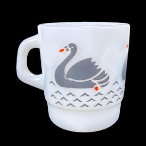 May include: White ceramic mug featuring a grey swan illustration with orange accents. The swan is depicted on water with grey wave-like patterns. The mug has a rounded handle and a stacked design.