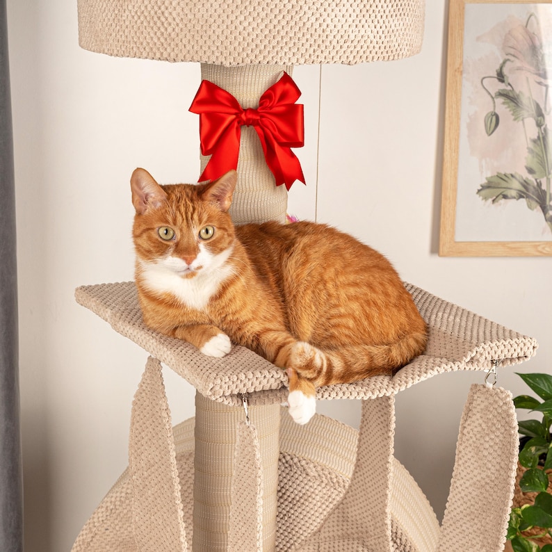 May include: A beige cat tree with a red bow on top. A ginger cat with white markings is lying on a platform on the cat tree. The cat is looking directly at the camera.