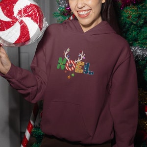 以下が含まれることがあります： クリスマスをテーマにしたデザインのマルーンのパーカー。カラフルな文字で「NOEL」と書いてあり、お祝いのアクセントが付けられています。文字はオーナメント、キャンディーケーン、トナカイの角で飾られています。