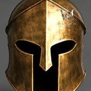 May include: A metallic gold Spartan-style helmet featuring a pointed crest, a central nasal guard, and wide eye openings. The surface has a weathered, antique finish with visible dark patina. The text WARRIOR is printed on the right side.