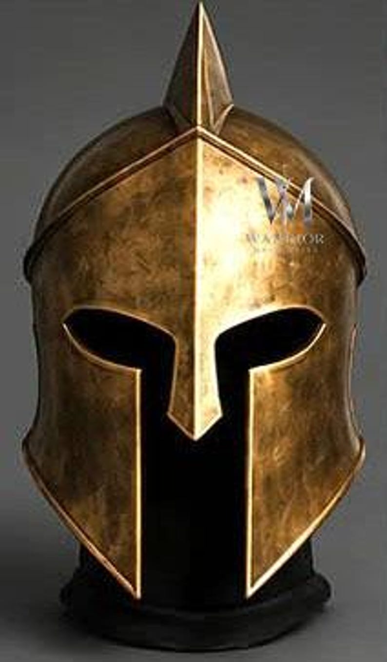 May include: A metallic gold Spartan-style helmet featuring a pointed crest, a central nasal guard, and wide eye openings. The surface has a weathered, antique finish with visible dark patina. The text WARRIOR is printed on the right side.