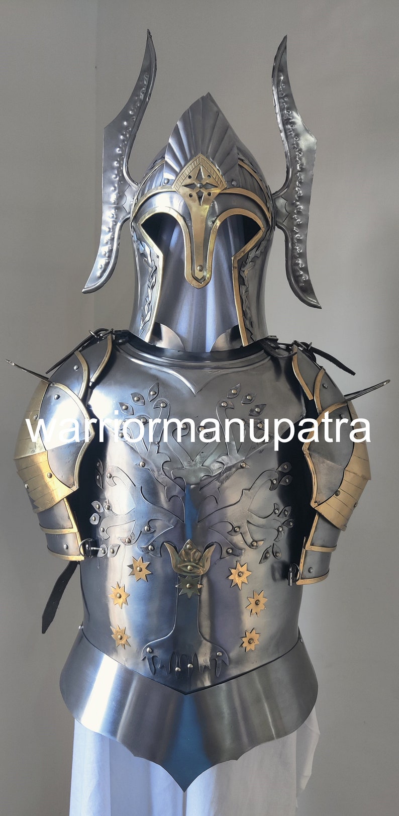 May include: A polished steel suit of armor featuring a helmet with two curved side fins and a brass-trimmed visor. The chest plate displays an engraved tree motif with brass star accents and the text warriormanupatra across the center.