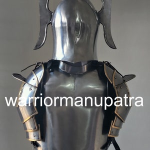 May include: A polished steel suit of armor featuring a pointed helmet with two curved, wing-like side projections, a breastplate, and articulated shoulder pauldrons with gold-toned trim. The text "warriormanupatra" is printed across the chest.