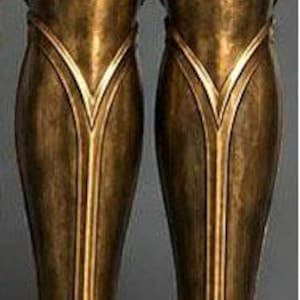 May include: A pair of metallic bronze greaves featuring a smooth, curved design with raised vertical ridges and a V-shaped accent near the top. The surface has a weathered, antiqued finish with subtle highlights and shadows across the contoured metal.