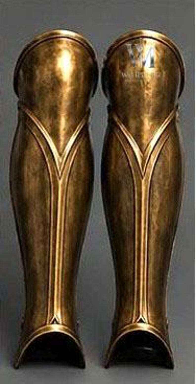 May include: A pair of metallic bronze greaves featuring a smooth, curved design with raised vertical ridges and a V-shaped accent near the top. The surface has a weathered, antiqued finish with subtle highlights and shadows across the contoured metal.