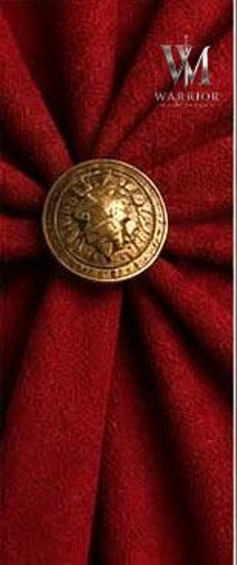 May include: A circular brass-toned metal button featuring a relief of a lion head, centered on a deep red fabric with radiating folds. The text "VM WARRIOR" is visible in the upper right corner, accompanied by a small sword icon above the letters.