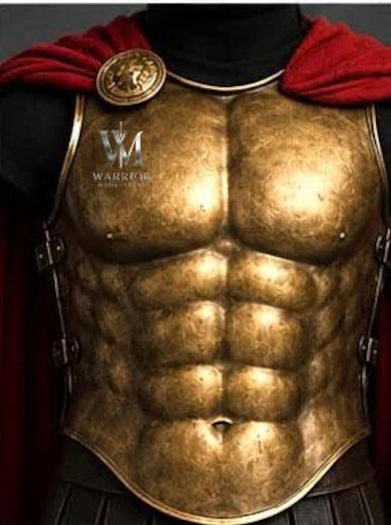 May include: A metallic gold-toned breastplate featuring a sculpted anatomical torso design with a defined six-pack. The armor includes a red fabric cape secured by a circular gold medallion and displays the text VM WARRIOR on the upper left chest.