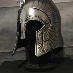 May include: A metallic silver helmet with a pointed crown and integrated cheek guards. The surface features embossed wing patterns on the sides and intricate scrollwork along the front edges. The logo text WARRIOR MANUPAL is visible.