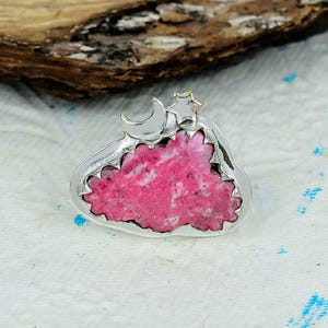 Peut inclure: Une bague en argent dotée d'un sertissage dentelé en forme de nuage contenant une pierre rose texturée et vive. Le haut de la monture en argent est orné d'un petit croissant de lune et d'une étoile à cinq branches, créant un élément céleste.