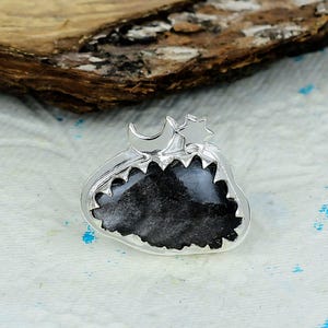 May include: A silver ring featuring a dark, textured, irregular black stone set within a jagged metal frame. The top of the setting is adorned with a small, polished silver crescent moon and a five-pointed star, creating a celestial design.