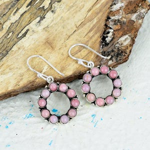 May include: A pair of silver-toned drop earrings featuring a circular frame set with small, smooth, round pink stones. Each earring hangs from a curved silver-toned hook, with the circular design composed of ten individual pink cabochons.