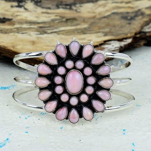 Peut inclure: Un bracelet manchette en argent avec un motif floral central. Le design est composé de nombreuses pierres roses en forme de goutte d'eau entourant une pierre ovale plus grande. Le bracelet a un design à trois bandes.