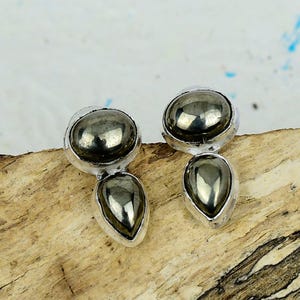 Può includere: Orecchini argentati con una pietra grigio scuro e metallica di forma rotonda, sormontata da una pietra a forma di goccia. Gli orecchini sono esposti su una superficie di legno. Le pietre hanno una lucentezza metallica.