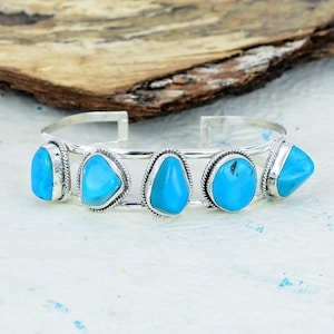 May include: A silver cuff bracelet with five bright turquoise stones. Each stone is uniquely shaped and set in a detailed silver bezel. The bracelet's open design allows for easy wear, showcasing the bright blue gemstones.