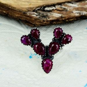 Pode incluir: Um anel em forma de V com cinco rubis vermelho-escuros facetados em forma de pera, cravados em prata de lei oxidada. As pedras estão dispostas simetricamente com um rubi central na base e dois pares de rubis estendendo-se para cima.