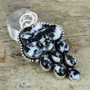 Peut inclure: Pendentif en argent orné d'une grande pierre en forme de nuage avec un motif noir et blanc, entourée de petites pierres en forme de goutte. Le pendentif a un bélière en argent et est posé sur une surface en bois.