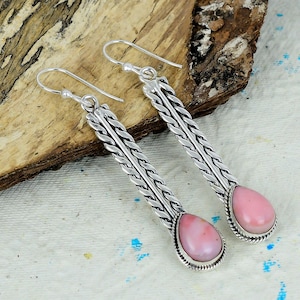 May include: A pair of silver earrings with a woven design. Each earring features a teardrop-shaped, light pink stone. The earrings have a hook closure and are displayed on a wooden surface.
