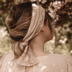 May include: A woman with brown hair wearing a light brown knitted headband with a delicate lace pattern. The headband is tied in a knot at the back of her neck.