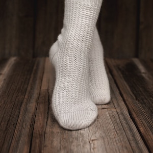 Könnte beinhalten: Ein Paar hellgraue Stricksocken. Die Socken haben einen gerippten Bund und ein strukturiertes Muster an Bein und Fuß. Die Socken befinden sich auf einer Holzoberfläche.