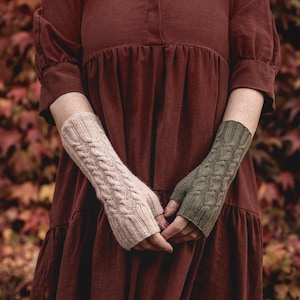 Könnte beinhalten: Ein Paar fingerlose Handschuhe mit Zopfmuster. Ein Handschuh ist hellbeige, der andere olivgrün. Die Handschuhe sind aus einem weichen, strukturierten Material gefertigt und sollen die Hände warm halten, während die Finger frei bleiben.