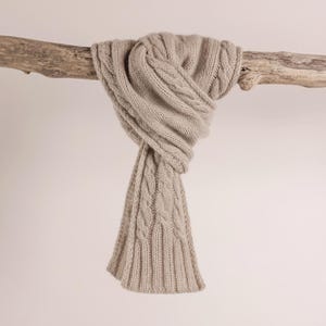 Peut inclure: Une écharpe beige en tricot torsadé, drapée sur un morceau de bois flotté. L'écharpe présente un motif tissé texturé et un bord côtelé. La couleur neutre et le design classique suggèrent un accessoire d'hiver douillet.