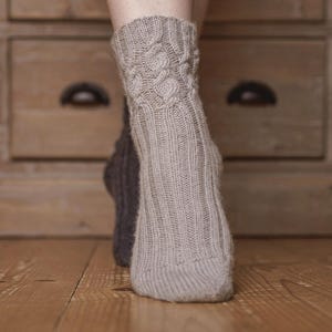 Könnte beinhalten: Nahaufnahme eines Paares gestrickter Socken. Die vordere Socke ist hellgrau mit einem Rippenmuster am Fuß und einem Zopfmuster am Bündchen. Die zweite Socke ist dunkler grau. Die Socken liegen auf einem Holzboden.