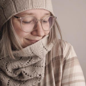 May include: A person wearing a beige knitted hat, scarf, and glasses. The scarf has a decorative openwork pattern. The person is also wearing a plaid shirt. The image is a close-up shot.
