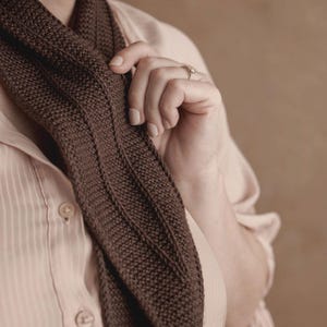 Könnte beinhalten: Ein dunkelbrauner Strickschal mit texturiertem Kraus-rechts-Muster und vertikalen Rippendetails, drapiert über einem hellbeigen Hemd mit Knopfleiste. Der Schal weist eine durchgehend gleichmäßige Strickkonstruktion in einem tiefen Erdfarbton auf.