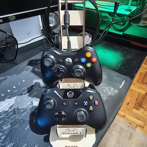 Könnte beinhalten: Ein Holzständer für Gamecontroller mit zwei schwarzen Xbox-Controllern und einem Headset. Der Ständer hat eine Basis mit dem Text "PAUSE, RECHARGE, PLAY." Die Controller sind gestapelt, der obere ist mit einem Kabel verbunden. Der Ständer steht auf einem Schreibtisch.