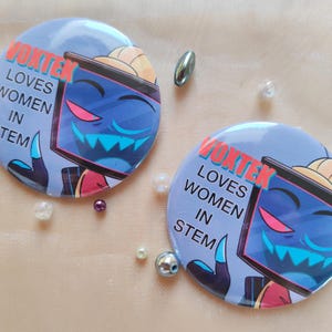 Op de afbeelding: Twee ronde buttons met de tekst "VOXTEK LOVES WOMEN IN STEM". De buttons tonen een blauw personage met een scherm als gezicht, een hoed en een hand. De achtergrond is lichtblauw. Decoratieve kralen omringen de buttons.